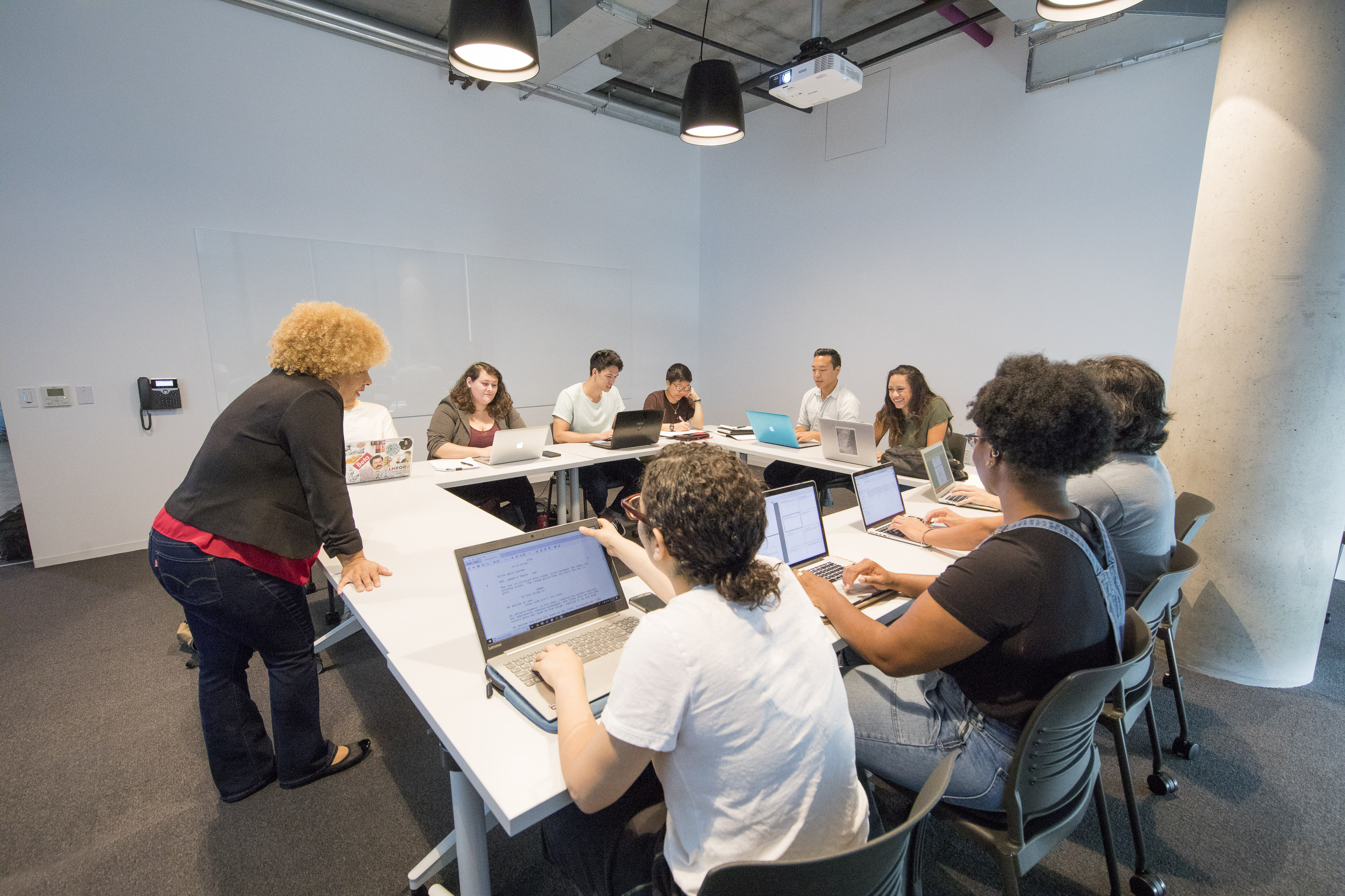 Professor stands at a square table in front of a group of students on their laptops